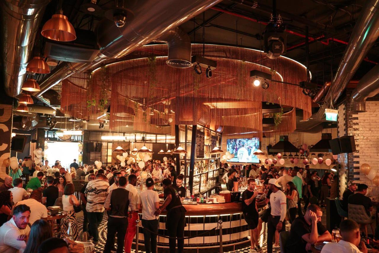 Five Jumeirah Village Dubai Ngoại thất bức ảnh The photo depicts a lively bar or restaurant scene filled with people socializing. The interior design features a modern aesthetic with a mix of industrial elements, such as exposed pipes and metallic accents. A large, circular bar is the focal point