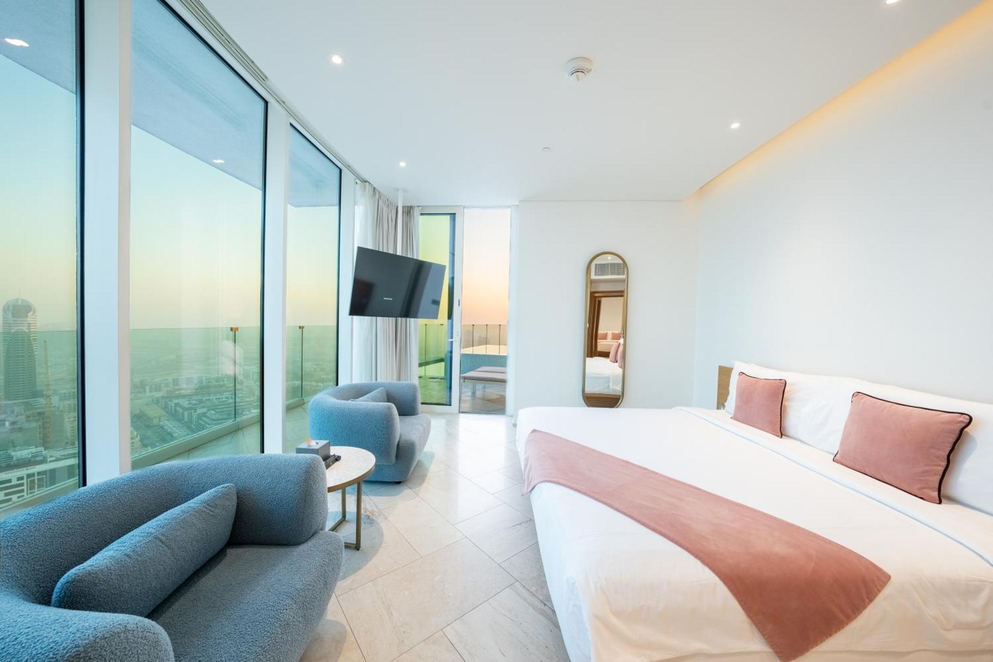 Five Jumeirah Village Dubai Ngoại thất bức ảnh The photo depicts a modern hotel room featuring large windows that provide a view of the outside. The room includes a spacious bed with a pink accent blanket, a cozy blue sofa, a small round table, and a wall-mounted television. There is also a mirro