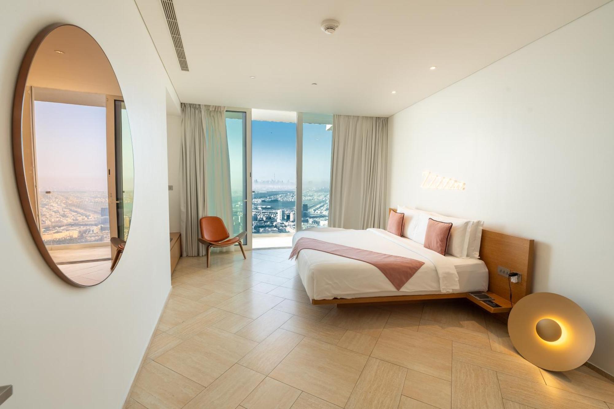 Five Jumeirah Village Dubai Ngoại thất bức ảnh The photo shows a modern bedroom with a minimalist design. There is a large bed with white bedding and pink accents, positioned against a wall. To the side, there is a round mirror and a stylish chair. The room features large windows dressed with lig