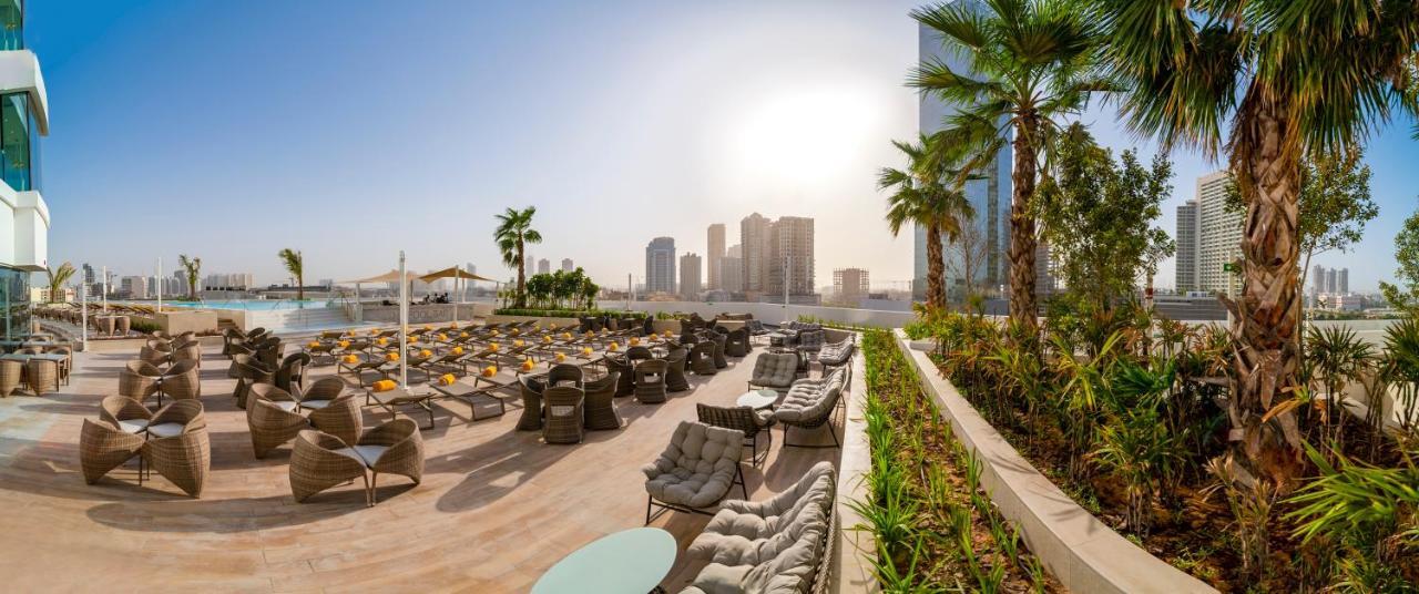 Five Jumeirah Village Dubai Ngoại thất bức ảnh The photo showcases a luxurious outdoor seating area, likely part of a resort or hotel. In the foreground, there are well-arranged lounge chairs and seating options, some with cushions, placed on a tiled surface. The area is bordered by lush green pl